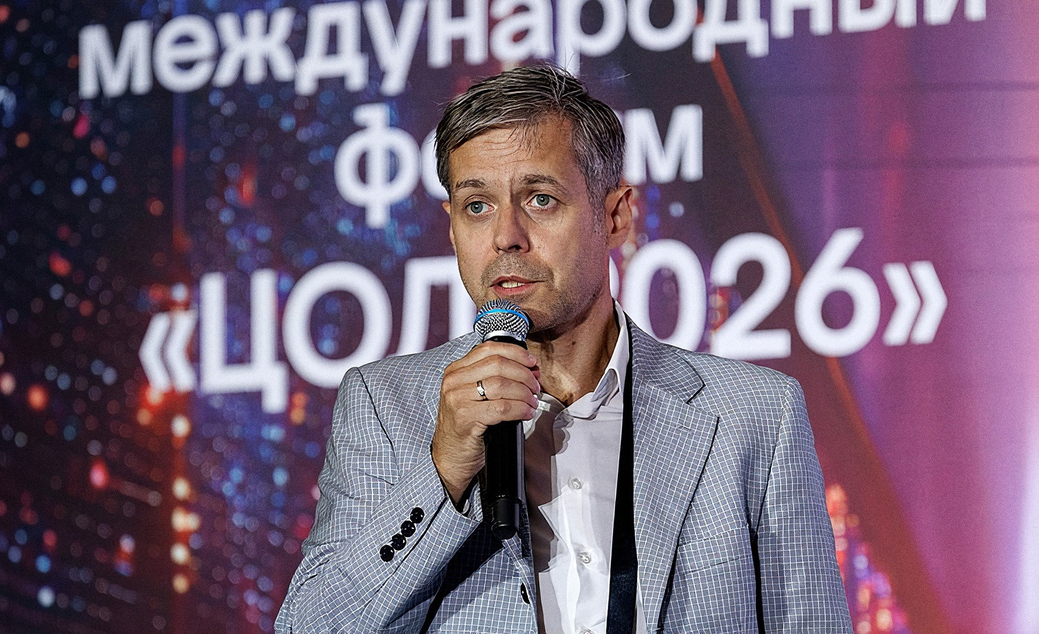 Спикер в светлом пиджаке говорит в микрофон на фоне заставки с названием форума