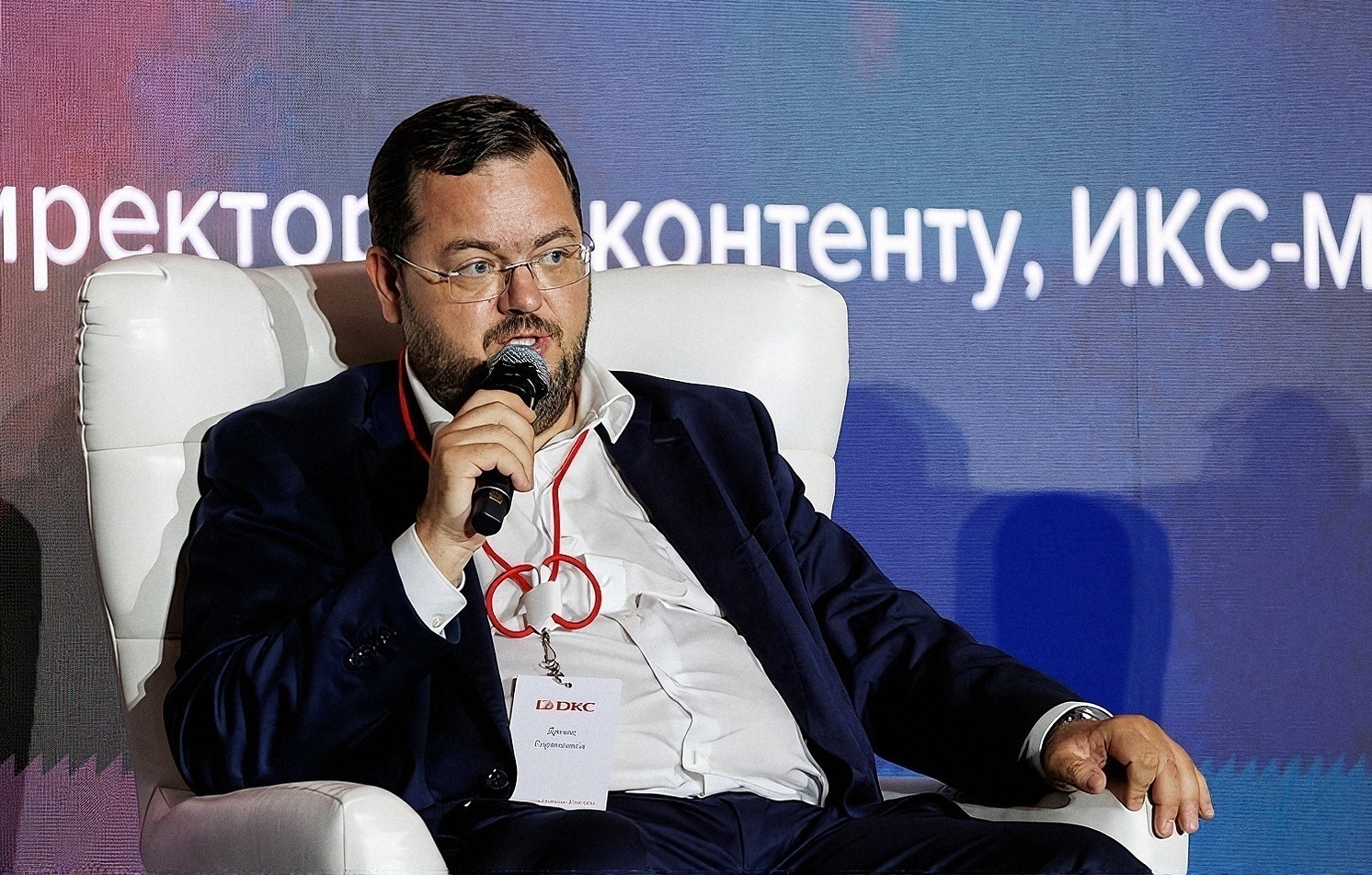 Участник дискуссии с бейджем DKC отвечает на вопрос, сидя в кресле на сцене