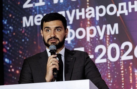 Докладчик в тёмном костюме у трибуны форума с микрофоном в руке