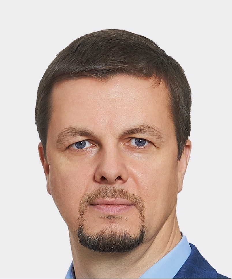 Мужчина с бородой и короткой стрижкой в тёмном костюме и голубой рубашке, лёгкая улыбка, светлый фон