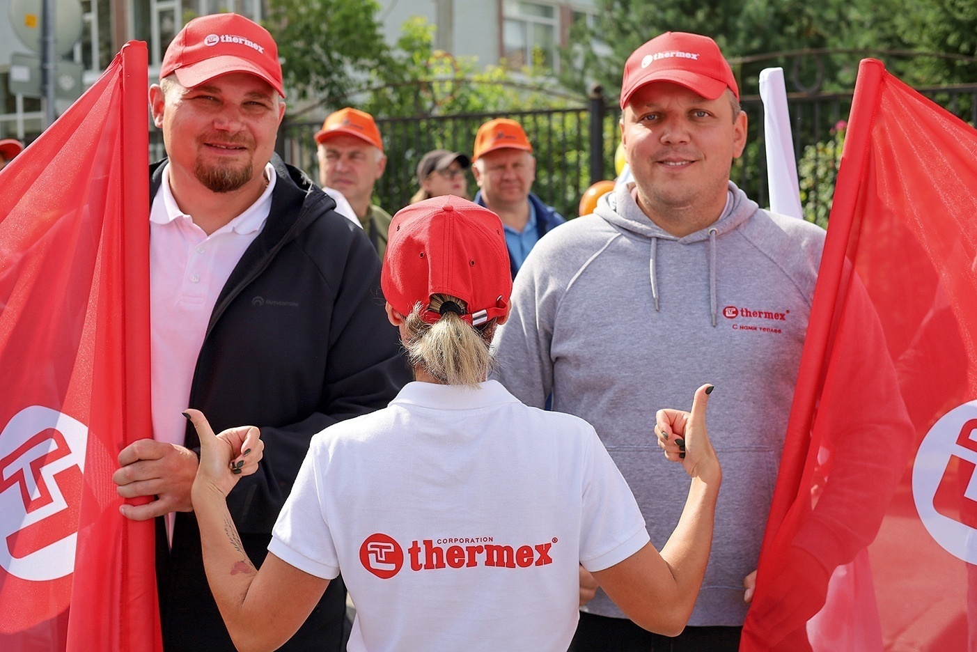 Сотрудники «Термекс» в красных кепках улыбаются и показывают большие пальцы на фоне фирменных флагов