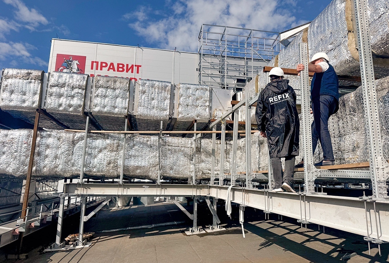 Рабочие в касках на стальной площадке кровли у многоярусной несущей конструкции с утеплённым воздуховодом на фоне неба